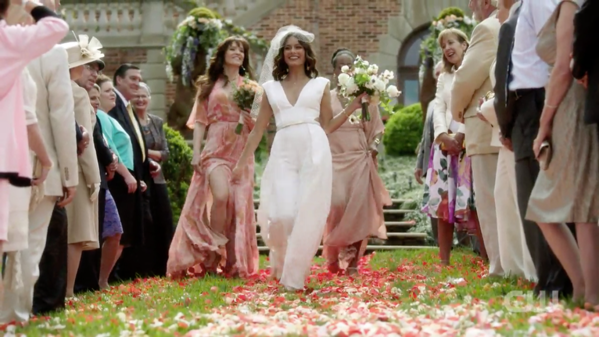 Woman in dumb white pantsuit (Cristal) swaying on a petal-strewn wedding aisle. Two bridesmaids in pale pink behind her.