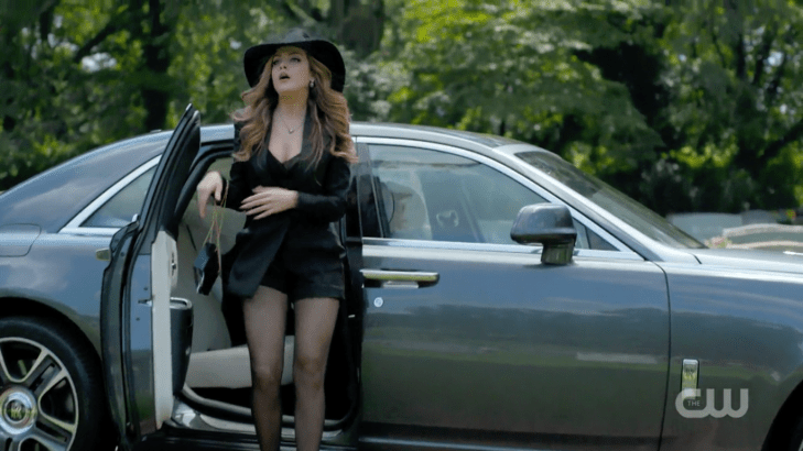 Fallon emerges from a car wearing black satin hot pants, a satin jacket, ab lack purse, a giant black hat, and sheer black tights.