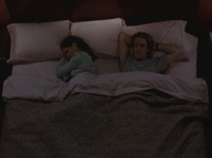 Joey and Dawson lie in a queen-sized bed, Joey facing away from Dawson on her side, Dawson with his hands clasped behind his head and his hair looking very ... tall.