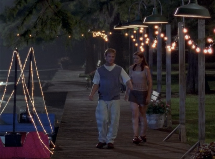 Joey and Dawson walk down a pier strewn with twinkle lights. Joey wears a pink tank top and a camo-green skirt, Dawson wears a giant shirt, a sweater vest, large white chinos, and a necklace.