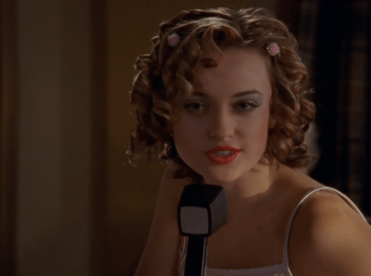 Abby wears corkscrew curls, pink pompom scrunchies, bright tomato-red lipstick.