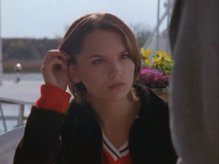 Devon (Rachael Leigh Cook) sitting outside by the water and some flowers, pushes hair behind ears and gives intense stare.