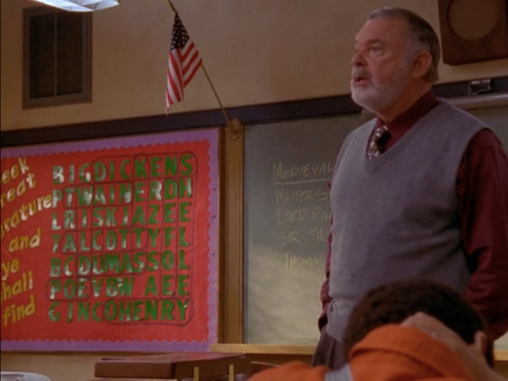 Teacher in sweater vest stands in front of display with American flag, chalkboard, and a word search that says "BIGDICKENS" across the top row.