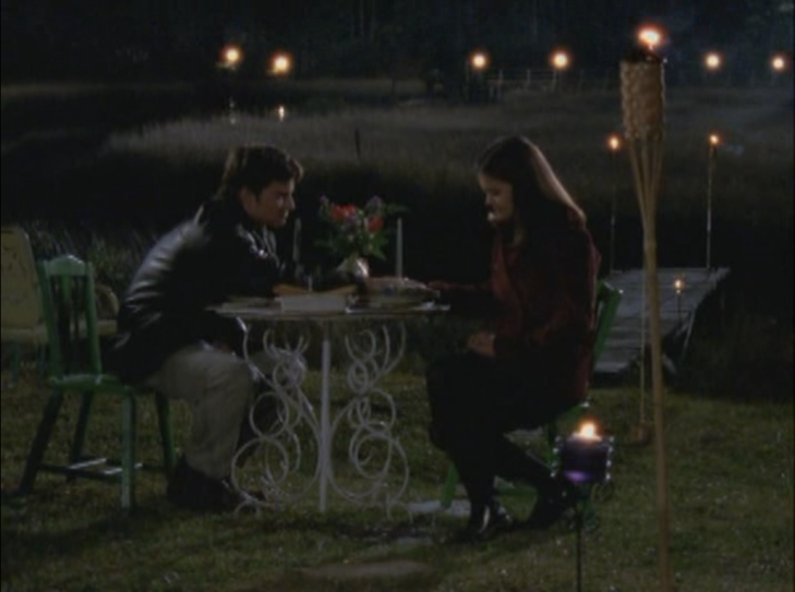 Joey and Jack sit at a table outdoors with flowers and candles in front of the creek. They are holding hands and look sad.