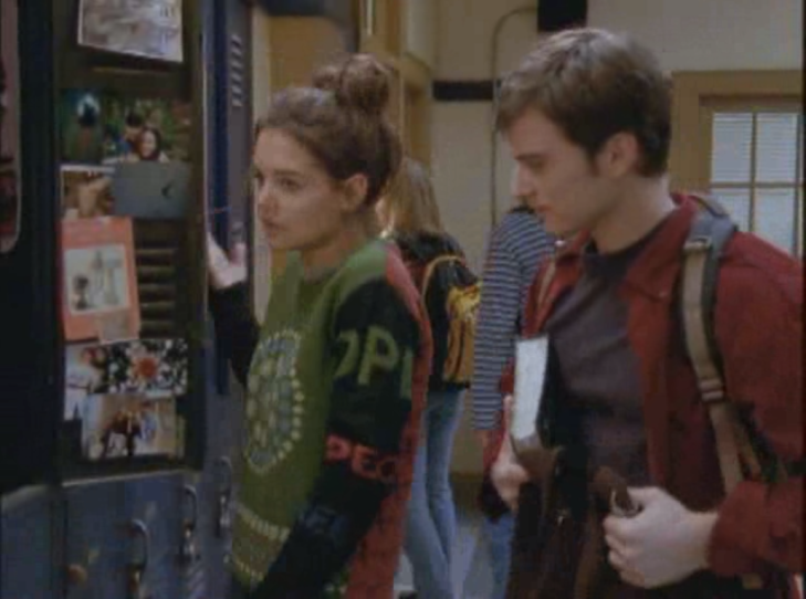 Joey standing at locker, in messy bun and a sweater with a green front, red back, black arms, w/ random words written on the arms and a circular mandala on the front.