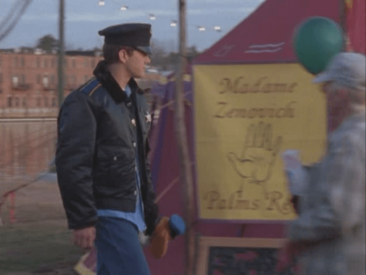 Pacey walks by the fortune-teller's sign in a black puffy jacket and a black officer's cap.