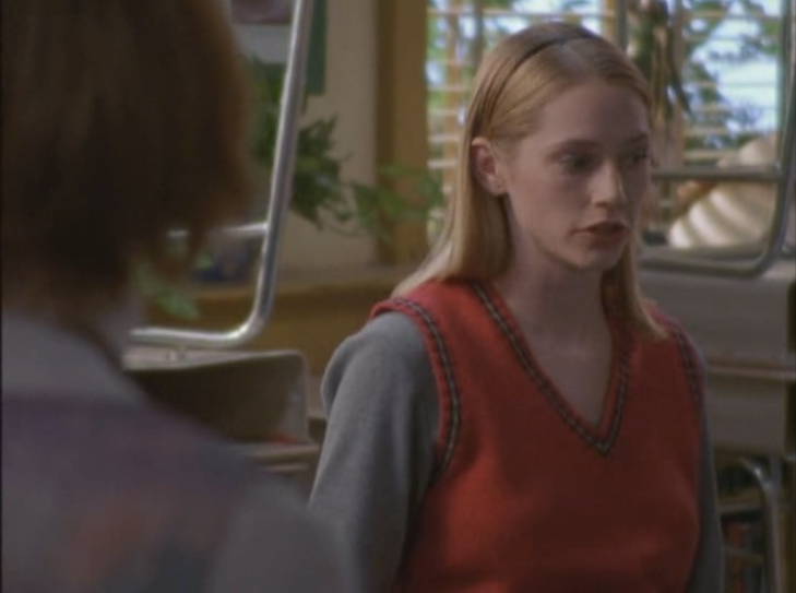 In a classroom, Andie wears a red embroidered sweater vest over a light blue sweater.