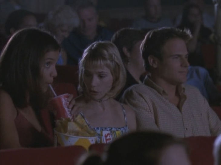 Movie theater. Joey sits left, sucking on a straw from a Coke cup, holding popcorn; Andie, middle, looks at her in annoyance, and Rob, right, faces forward with a smirk.