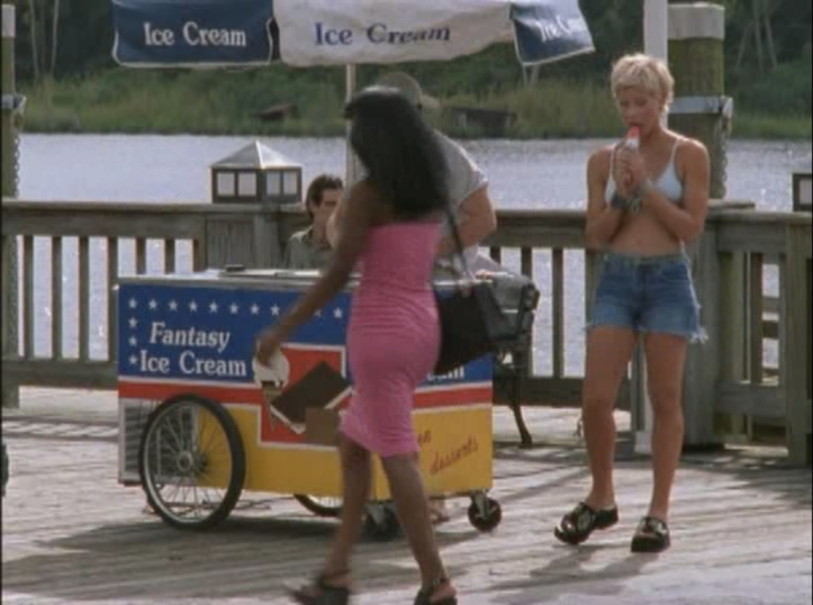 Eve eats a popsicle on the dock next to an ice cream cart, wearing a bikini top and jean short-shorts.