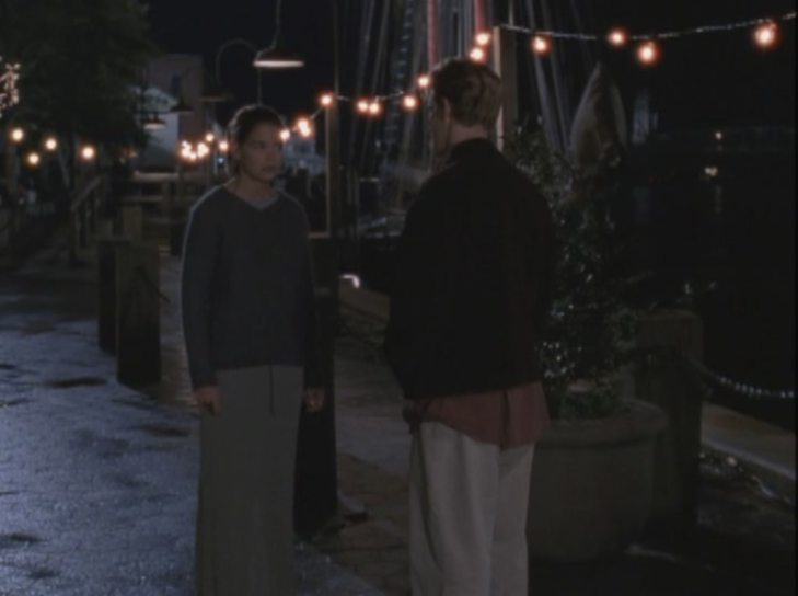 Joey and Dawson face each other on the dock, strung with twinkle lights. Joey wears a loose long sleeved sweater whose neckline barely goes below her collarbone in a dark grey-blue, and a long shapeless skirt in dark grey-green.