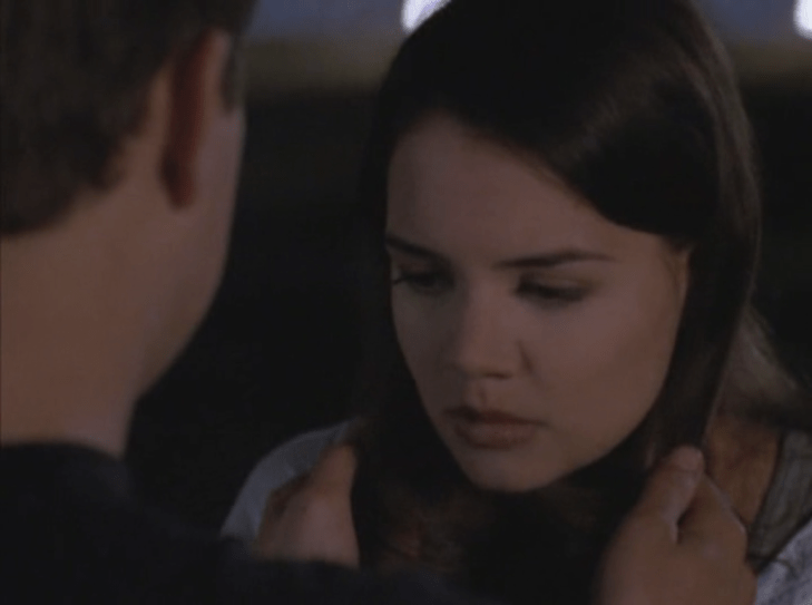 Joey looks down, looking sad, and Pacey (facing away from camera) tugs gently on her hair.