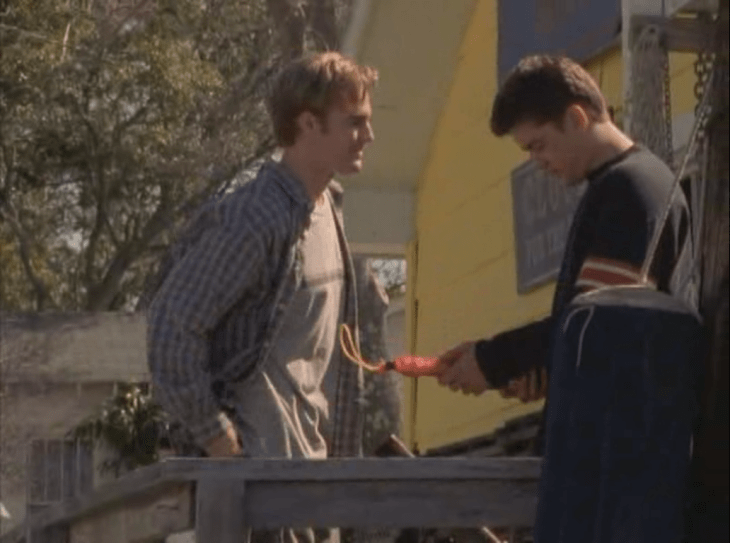 Dawson hands Pacey a champagne bottle with a loop of rope coming out of it.