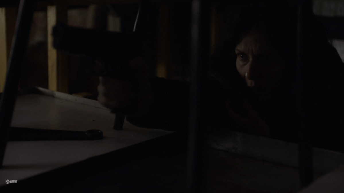 A dark room, with Carrie in a brunette wig kneeling and pointing a gun in front of her.