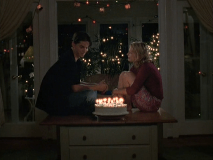 Interior: in front of a bunch of twinkle lights, Jen and Drue sit facing each other on a table, with a birthday cake fully lit with candles between them.