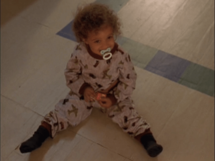 A little boy in a onesie with a pacifier, a big fro, and an adorable smile sits on the floor. (Shot from above, interior.)