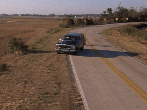 A car pulls off the side of the road. Fields are seen on the left side, and yellow signs warning of a curve in the road behind.