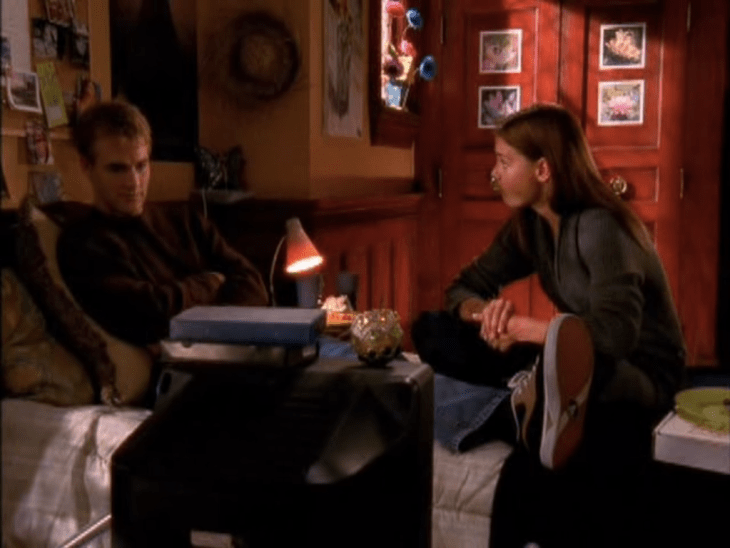 Joey and Dawson sit awkwardly on the dorm bed, with a TV in front of them. Joey is cross-legged, Dawson stretched out with his arms crossed.