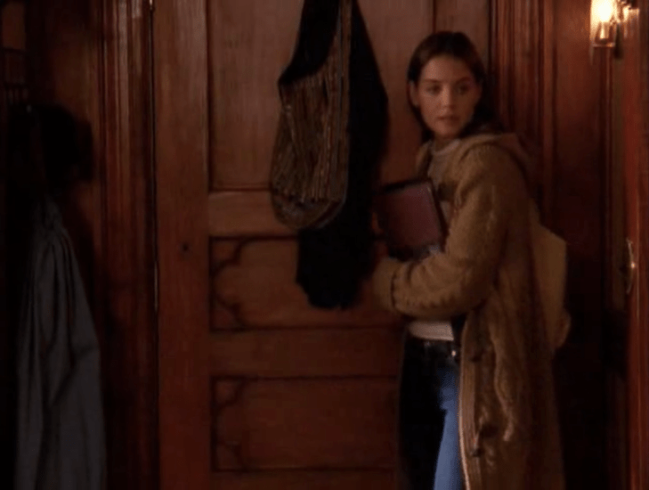 Joey closes the door of her dorm. She is wearing a camel-colored, long, thick sweater coat with jeans and carrying books.