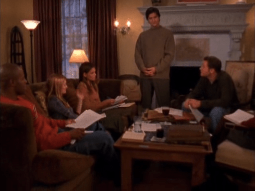 Some students (three white - one of them is Joey and one black) sit around a coffee table with papers scattered everywhere. An older brunette man (Professor Wilder) stands at the side of the room, clasping his hands.