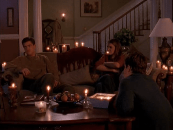 Long shot of three people - two brunette white men (Jack, at the coffee table; Pacey, on the couch on the left) one brunette white woman (Joey, on the couch, right) -- in a living room surrounded by candles.