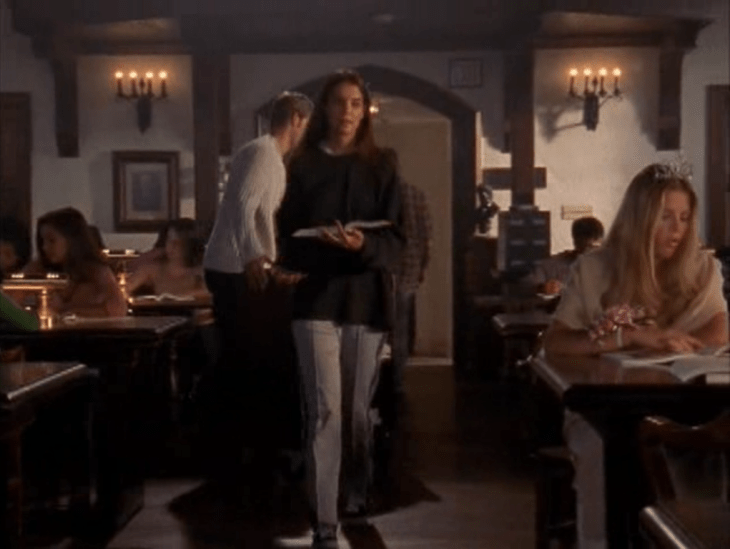 Joey (brunette white woman) walks through the library past a blonde white woman (Audrey) in a tiara. She is wearing gray sweatpants that are pleated.