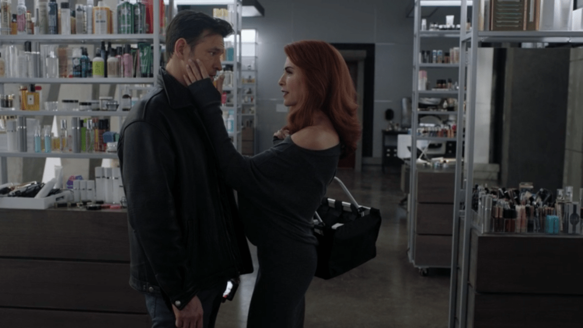 Inside a room filled with beauty products, a white woman with red hair and a fancy dress (Kitty) holds her hand up to the face of a white man with brown hair and a jacket (Dominic).