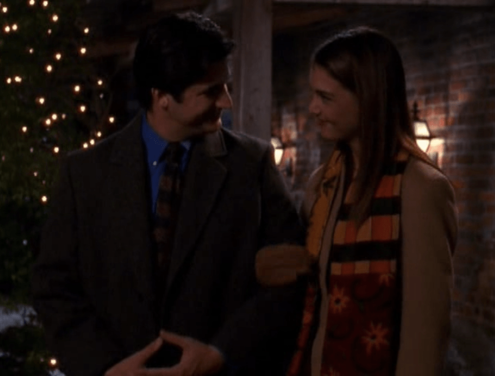 Professor Wilder, wearing a coat and tie, holds out his elbow and Joey holds it in her hand, wearing a winter jacket, scarf, and gloves. Both are looking at each other and smiling.