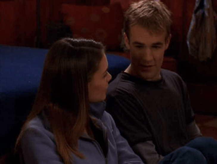 A young white woman and a young white man (Joey and Dawson) sit against a bed, shoulders touching.