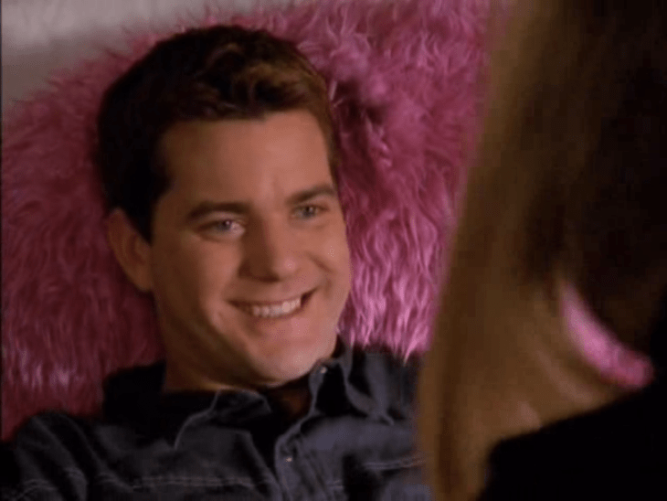 A white brunette man (Pacey) is seen grinning, resting his head on a pink feathery pillow.
