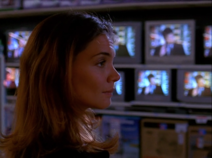 Interior - Joey, seen from the side, looks like she's about to smile, while a lot of small TVs are arranged behind her with blurry