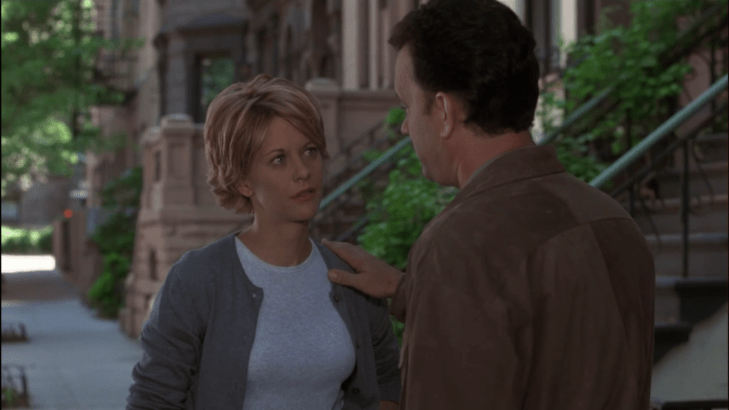 Kathleen (facing camera, in a sweater suit) and Joe (facing away) stand on a street covered in spring greenery; he has his hand on his shoulder, and she looks sad.