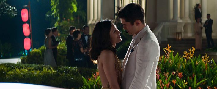 Outside in a lush garden outside a large house, a young woman in a formal dress stares lovingly up at a handsome man in a dinner jacket.
