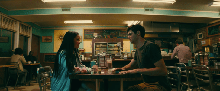 A teenaged girl and boy sit at a nearly-empty diner, facing each other across the table.