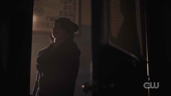 Veronica stands in a doorway with a dramatic black hat and a dramatic black coat.