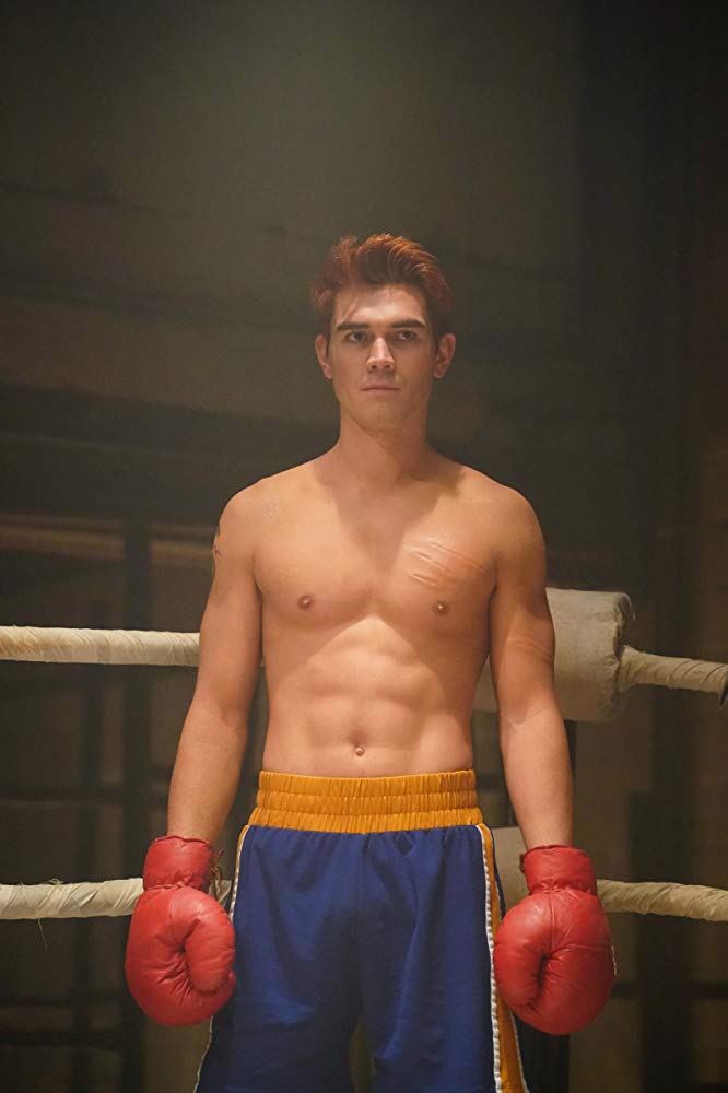 Archie, shirtless, standsin a boxing ring wearing giant red gloves and blue shorts.