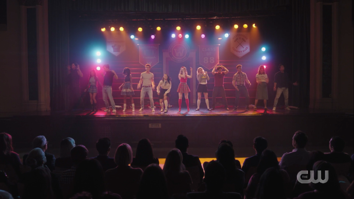 The cast stands on a lit stage in 80s clothes, dancing.