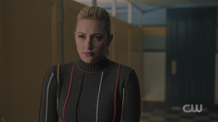 Betty stands in a bathroom, wearing a super ugly sweater (gray, with yellow, pink, black, and white yarn cylinders glued onto it)