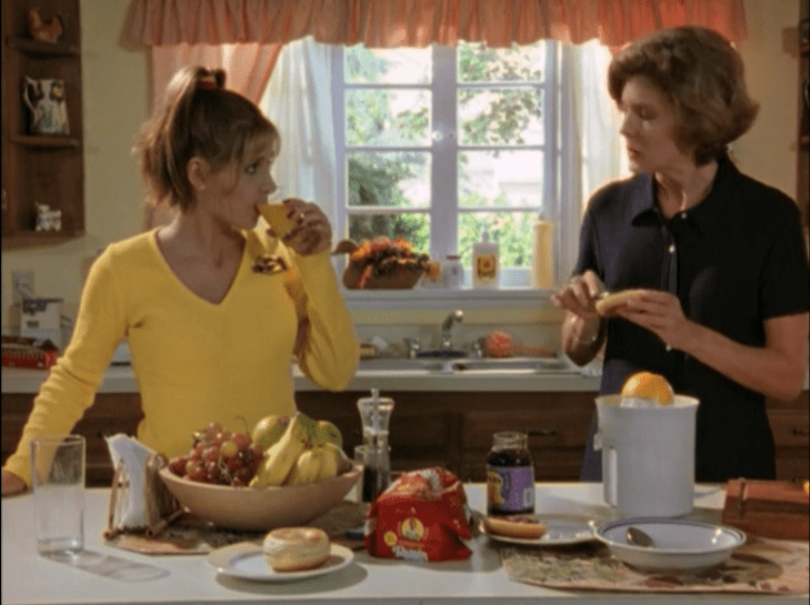 Buffy wears a cheerleading outfit and drinks juice while in the kitchen with her mom.