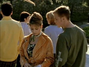 Surrounded by kids walking away from her on an outdoor path, Buffy looks down. She is wearing an orange velvet belted trench, and a teeny tiny green scarf.