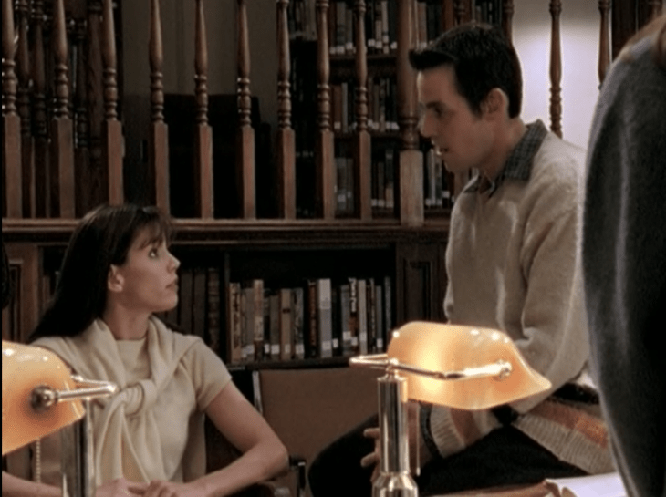 Cordelia and Xander in the library. Cordelia is sitting, but her high-necked yellow shirt and yellow cardigan tied around her shoulders are visible.