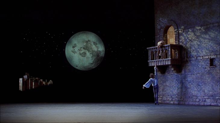 Image description: Onstage, a man on tiptoe reaches up to a woman on a balcony.