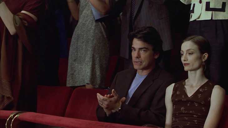 Image description: In an auditorium, Jonathan talks out of the side of his mouth to Kathleen, hands mid-unenthusiastic-clap. They are seated, everyone else is standing