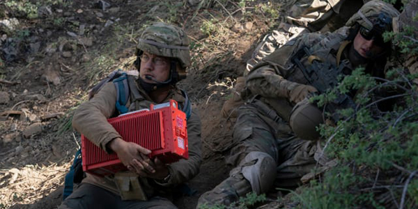 Max clutches a red box, in camo gear, next to another soldier.